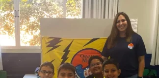 Professora posa com alunos em frente a uma maquete escolar, representando o processo seletivo emergencial na Educação de Florianópolis.