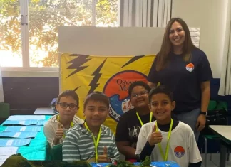 Professora posa com alunos em frente a uma maquete escolar, representando o processo seletivo emergencial na Educação de Florianópolis.