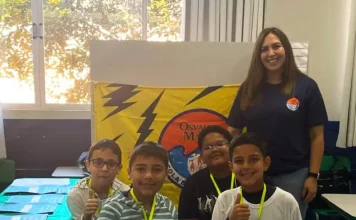 Professora posa com alunos em frente a uma maquete escolar, representando o processo seletivo emergencial na Educação de Florianópolis.