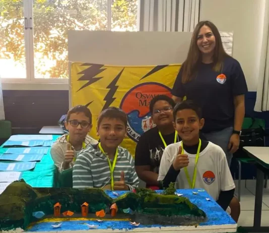 Professora posa com alunos em frente a uma maquete escolar, representando o processo seletivo emergencial na Educação de Florianópolis.