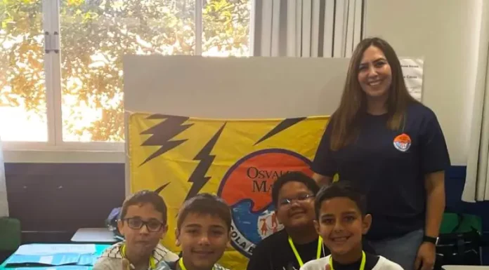 Professora posa com alunos em frente a uma maquete escolar, representando o processo seletivo emergencial na Educação de Florianópolis.