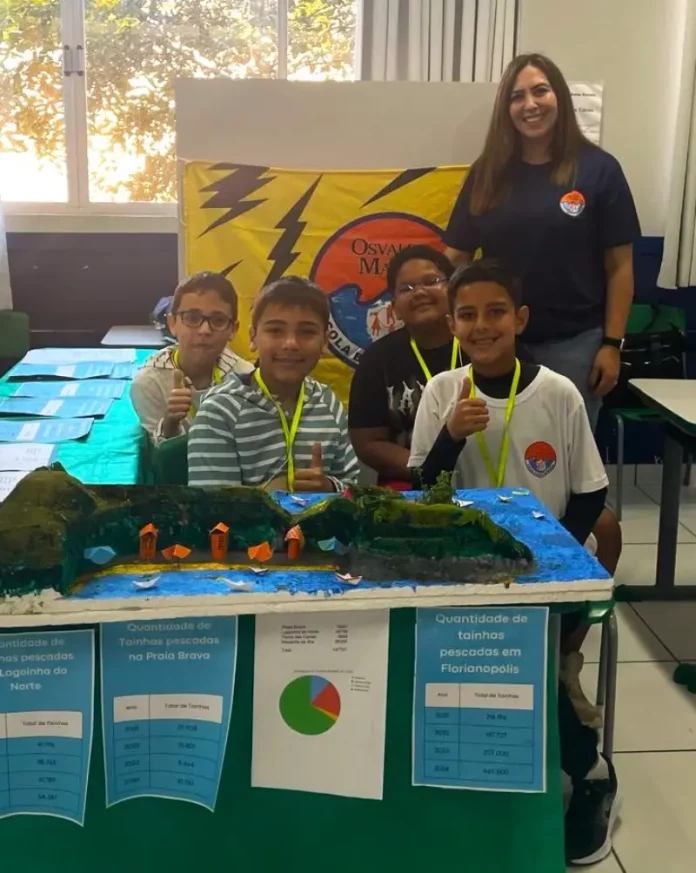 Professora posa com alunos em frente a uma maquete escolar, representando o processo seletivo emergencial na Educação de Florianópolis.