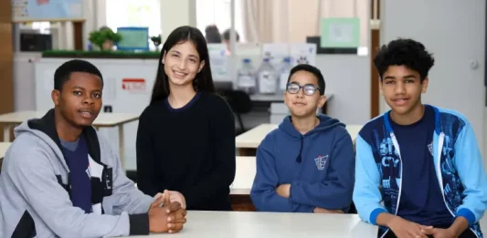 Estudantes migrantes — três meninos e uma menina — sentados juntos em uma mesa de sala de aula