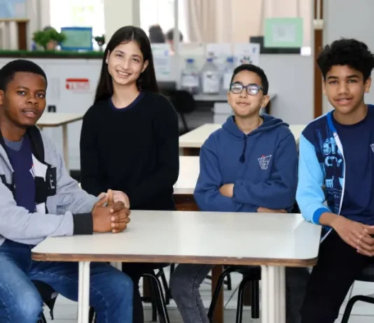 Estudantes migrantes — três meninos e uma menina — sentados juntos em uma mesa de sala de aula