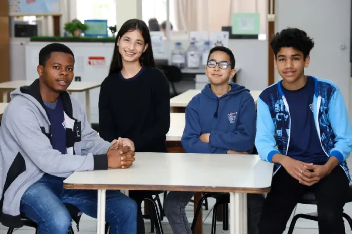 Estudantes migrantes — três meninos e uma menina — sentados juntos em uma mesa de sala de aula