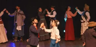 Integrantes da categoria adulta de danças tradicionais do Grupo Querência Açoriana se apresentam no palco durante a Fegasc, vestindo trajes típicos e executando coreografia gaúcha.