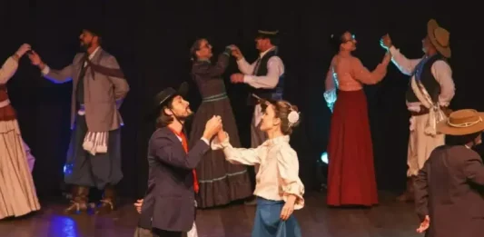 Integrantes da categoria adulta de danças tradicionais do Grupo Querência Açoriana se apresentam no palco durante a Fegasc, vestindo trajes típicos e executando coreografia gaúcha.