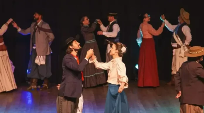Integrantes da categoria adulta de danças tradicionais do Grupo Querência Açoriana se apresentam no palco durante a Fegasc, vestindo trajes típicos e executando coreografia gaúcha.