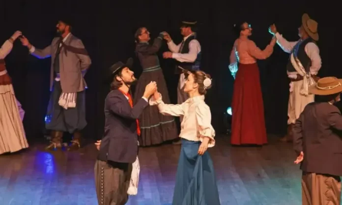Integrantes da categoria adulta de danças tradicionais do Grupo Querência Açoriana se apresentam no palco durante a Fegasc, vestindo trajes típicos e executando coreografia gaúcha.