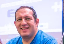 Cartunista Rodrigo Tramonte, vestindo camiseta azul com a estampa da capa de Humor Azul, posa ao lado do livro exposto, que aborda a experiência de autista no mercado de trabalho.