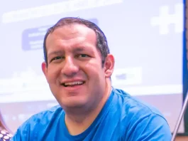 Cartunista Rodrigo Tramonte, vestindo camiseta azul com a estampa da capa de Humor Azul, posa ao lado do livro exposto, que aborda a experiência de autista no mercado de trabalho.