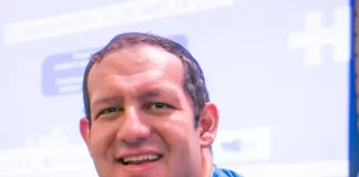 Cartunista Rodrigo Tramonte, vestindo camiseta azul com a estampa da capa de Humor Azul, posa ao lado do livro exposto, que aborda a experiência de autista no mercado de trabalho.