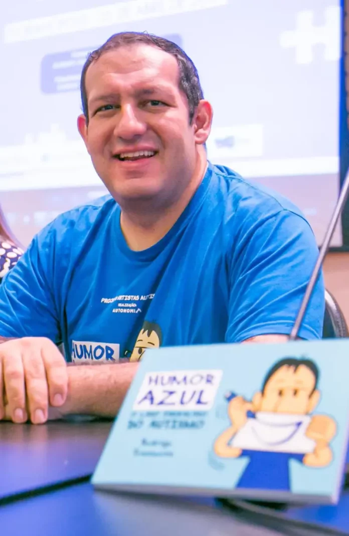 Cartunista Rodrigo Tramonte, vestindo camiseta azul com a estampa da capa de Humor Azul, posa ao lado do livro exposto, que aborda a experiência de autista no mercado de trabalho.