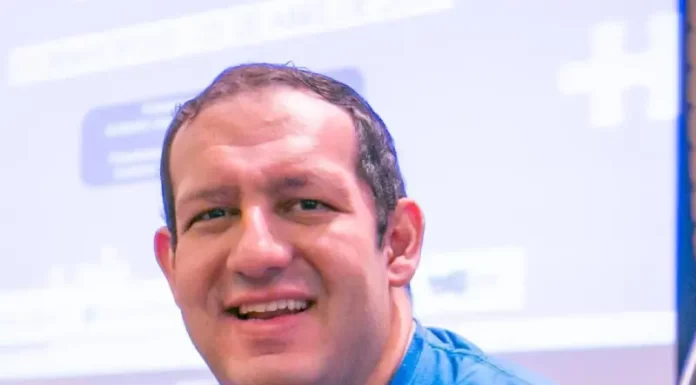 Cartunista Rodrigo Tramonte, vestindo camiseta azul com a estampa da capa de Humor Azul, posa ao lado do livro exposto, que aborda a experiência de autista no mercado de trabalho.