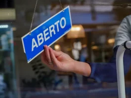 Mão de uma mulher segurando uma placa azul com a palavra “ABERTO” em branco, simbolizando a aprovação do Projeto de Lei que simplifica a abertura de empresas pela Alesc.