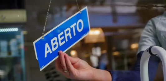 Mão de uma mulher segurando uma placa azul com a palavra “ABERTO” em branco, simbolizando a aprovação do Projeto de Lei que simplifica a abertura de empresas pela Alesc.