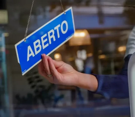 Mão de uma mulher segurando uma placa azul com a palavra “ABERTO” em branco, simbolizando a aprovação do Projeto de Lei que simplifica a abertura de empresas pela Alesc.