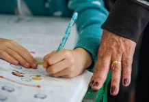 Pessoa adulta acompanha criança durante atividade de escrita, simbolizando ações de combate ao analfabetismo e incentivo à alfabetização na educação básica.