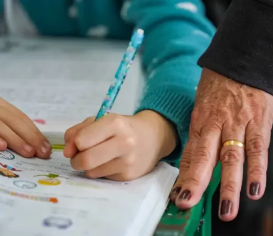 Pessoa adulta acompanha criança durante atividade de escrita, simbolizando ações de combate ao analfabetismo e incentivo à alfabetização na educação básica.