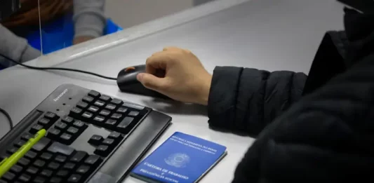 Santa Catarina acumula 83,8 mil novas vagas de empregos formais em 2025 Braço de um homem usando o mouse ao lado de um teclado, com uma carteira de trabalho sobre a mesa, ilustrando o registro e controle de empregos formais em Santa Catarina.