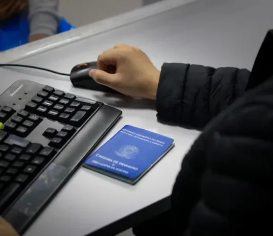 Santa Catarina acumula 83,8 mil novas vagas de empregos formais em 2025 Braço de um homem usando o mouse ao lado de um teclado, com uma carteira de trabalho sobre a mesa, ilustrando o registro e controle de empregos formais em Santa Catarina.