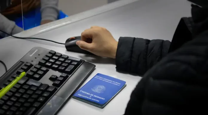 Santa Catarina acumula 83,8 mil novas vagas de empregos formais em 2025 Braço de um homem usando o mouse ao lado de um teclado, com uma carteira de trabalho sobre a mesa, ilustrando o registro e controle de empregos formais em Santa Catarina.