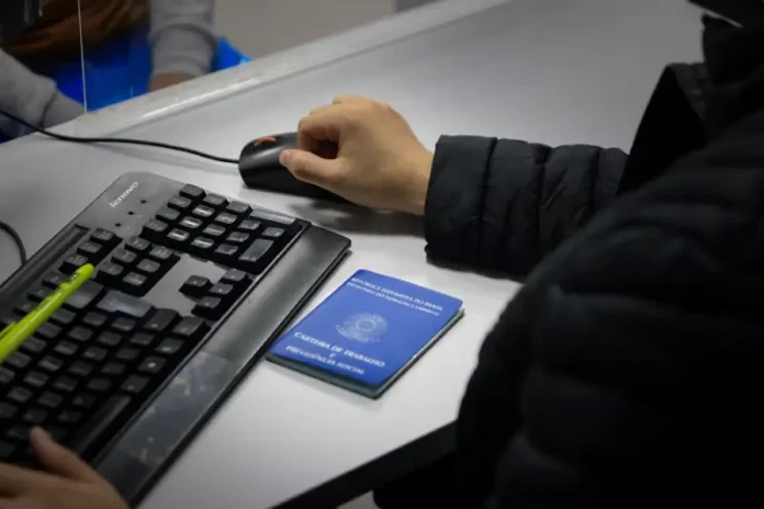 Braço de um homem usando o mouse ao lado de um teclado, com uma carteira de trabalho sobre a mesa, ilustrando o registro e controle de empregos formais em Santa Catarina.
