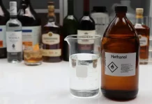 Mesa exibe diversas garrafas de bebidas alcoólicas ao lado de um frasco de metanol, ilustrando o alerta do Procon/SC sobre riscos de adulteração e intoxicação por metanol.