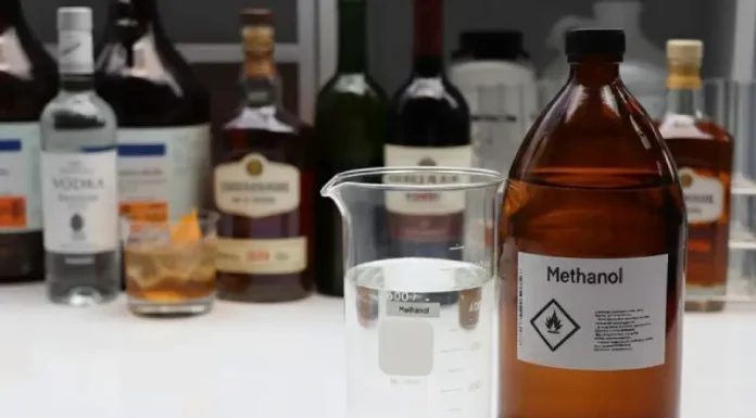 Procon/SC alerta sobre risco de bebidas adulteradas com metanol Mesa exibe diversas garrafas de bebidas alcoólicas ao lado de um frasco de metanol, ilustrando o alerta do Procon/SC sobre riscos de adulteração e intoxicação por metanol.
