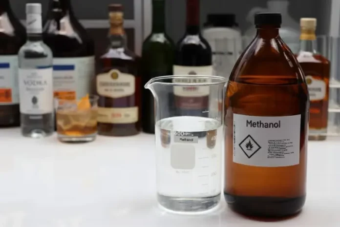 Mesa exibe diversas garrafas de bebidas alcoólicas ao lado de um frasco de metanol, ilustrando o alerta do Procon/SC sobre riscos de adulteração e intoxicação por metanol.