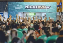 Imagem mostra a largada da corrida noturna 5K Platz Night Run, com uma multidão de participantes vestidos com camisetas verdes, braços erguidos, prontos para iniciar a prova. Ao fundo, há uma faixa com o nome do evento e um cronômetro zerado, indicando o início iminente da corrida. A foto ilustra a energia e a mobilização do público no evento esportivo realizado à noite em São José.