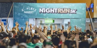 Imagem mostra a largada da corrida noturna 5K Platz Night Run, com uma multidão de participantes vestidos com camisetas verdes, braços erguidos, prontos para iniciar a prova. Ao fundo, há uma faixa com o nome do evento e um cronômetro zerado, indicando o início iminente da corrida. A foto ilustra a energia e a mobilização do público no evento esportivo realizado à noite em São José.
