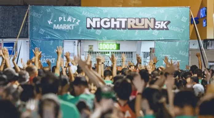 Imagem mostra a largada da corrida noturna 5K Platz Night Run, com uma multidão de participantes vestidos com camisetas verdes, braços erguidos, prontos para iniciar a prova. Ao fundo, há uma faixa com o nome do evento e um cronômetro zerado, indicando o início iminente da corrida. A foto ilustra a energia e a mobilização do público no evento esportivo realizado à noite em São José.