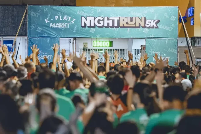 Imagem mostra a largada da corrida noturna 5K Platz Night Run, com uma multidão de participantes vestidos com camisetas verdes, braços erguidos, prontos para iniciar a prova. Ao fundo, há uma faixa com o nome do evento e um cronômetro zerado, indicando o início iminente da corrida. A foto ilustra a energia e a mobilização do público no evento esportivo realizado à noite em São José.