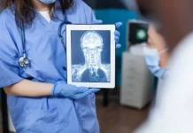 Profissional da saúde, usando jaleco e luvas, segura um tablet que exibe uma imagem de ressonância magnética do crânio e da coluna cervical. A imagem ilustra o uso da tecnologia digital e da inteligência artificial no diagnóstico por imagem, tema central de debates sobre inovação médica.