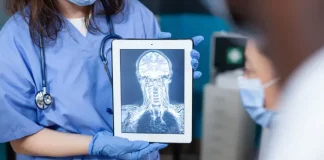 Profissional da saúde, usando jaleco e luvas, segura um tablet que exibe uma imagem de ressonância magnética do crânio e da coluna cervical. A imagem ilustra o uso da tecnologia digital e da inteligência artificial no diagnóstico por imagem, tema central de debates sobre inovação médica.