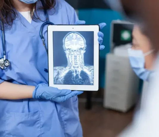 Profissional da saúde, usando jaleco e luvas, segura um tablet que exibe uma imagem de ressonância magnética do crânio e da coluna cervical. A imagem ilustra o uso da tecnologia digital e da inteligência artificial no diagnóstico por imagem, tema central de debates sobre inovação médica.