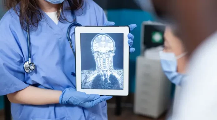 Profissional da saúde, usando jaleco e luvas, segura um tablet que exibe uma imagem de ressonância magnética do crânio e da coluna cervical. A imagem ilustra o uso da tecnologia digital e da inteligência artificial no diagnóstico por imagem, tema central de debates sobre inovação médica.