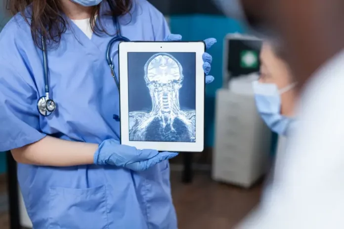 Profissional da saúde, usando jaleco e luvas, segura um tablet que exibe uma imagem de ressonância magnética do crânio e da coluna cervical. A imagem ilustra o uso da tecnologia digital e da inteligência artificial no diagnóstico por imagem, tema central de debates sobre inovação médica.