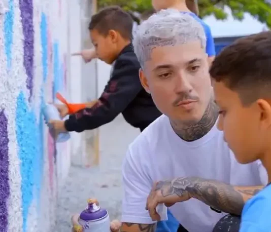 Rhuan Santo orienta uma criança sobre técnicas de grafite em parede, simbolizando a integração entre arte urbana e educação.