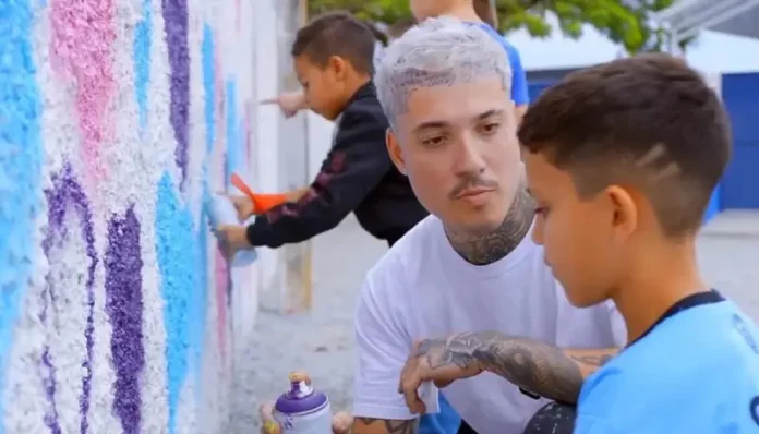 Rhuan Santo orienta uma criança sobre técnicas de grafite em parede, simbolizando a integração entre arte urbana e educação.