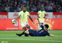 Jogador da seleção brasileira tenta avançar com a bola enquanto é desarmado por um defensor da seleção japonesa durante uma partida de futebol. A imagem ilustra o confronto entre Brasil e Japão, usada para representar o tema do desempenho da seleção brasileira.
