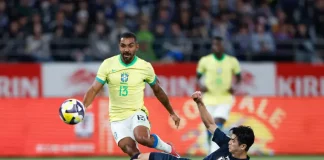 Jogador da seleção brasileira tenta avançar com a bola enquanto é desarmado por um defensor da seleção japonesa durante uma partida de futebol. A imagem ilustra o confronto entre Brasil e Japão, usada para representar o tema do desempenho da seleção brasileira.