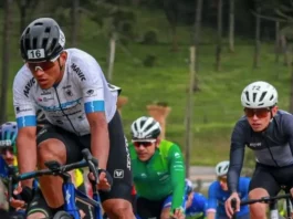 Grupo de ciclistas participa de uma competição em estrada durante o dia. Os atletas usam uniformes e capacetes de segurança, pedalando em alta velocidade. A imagem representa o contexto esportivo da Volta Ciclística de Santa Catarina, com foco no esforço físico dos competidores e no dinamismo da prova. Ao fundo, é possível ver uma área verde com árvores, sugerindo um percurso em meio à natureza.