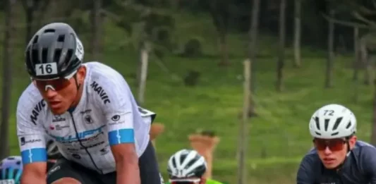 Grupo de ciclistas participa de uma competição em estrada durante o dia. Os atletas usam uniformes e capacetes de segurança, pedalando em alta velocidade. A imagem representa o contexto esportivo da Volta Ciclística de Santa Catarina, com foco no esforço físico dos competidores e no dinamismo da prova. Ao fundo, é possível ver uma área verde com árvores, sugerindo um percurso em meio à natureza.