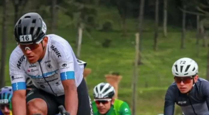 Grupo de ciclistas participa de uma competição em estrada durante o dia. Os atletas usam uniformes e capacetes de segurança, pedalando em alta velocidade. A imagem representa o contexto esportivo da Volta Ciclística de Santa Catarina, com foco no esforço físico dos competidores e no dinamismo da prova. Ao fundo, é possível ver uma área verde com árvores, sugerindo um percurso em meio à natureza.