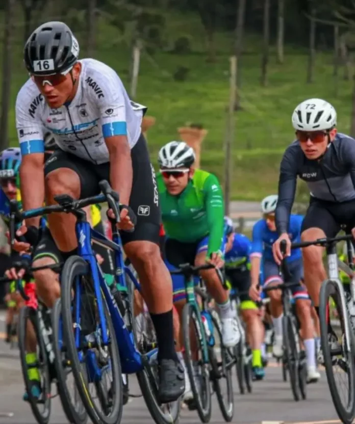 Grupo de ciclistas participa de uma competição em estrada durante o dia. Os atletas usam uniformes e capacetes de segurança, pedalando em alta velocidade. A imagem representa o contexto esportivo da Volta Ciclística de Santa Catarina, com foco no esforço físico dos competidores e no dinamismo da prova. Ao fundo, é possível ver uma área verde com árvores, sugerindo um percurso em meio à natureza.