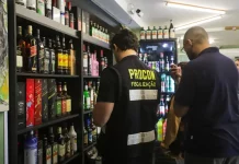 Agente do Procon de Florianópolis realiza fiscalização em bar da capital, observando prateleiras com garrafas de bebidas alcoólicas. A ação visa prevenir a venda de produtos adulterados, especialmente frente aos casos recentes de intoxicação por metanol.