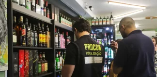 Agente do Procon de Florianópolis realiza fiscalização em bar da capital, observando prateleiras com garrafas de bebidas alcoólicas. A ação visa prevenir a venda de produtos adulterados, especialmente frente aos casos recentes de intoxicação por metanol.