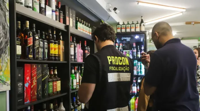 Agente do Procon de Florianópolis realiza fiscalização em bar da capital, observando prateleiras com garrafas de bebidas alcoólicas. A ação visa prevenir a venda de produtos adulterados, especialmente frente aos casos recentes de intoxicação por metanol.